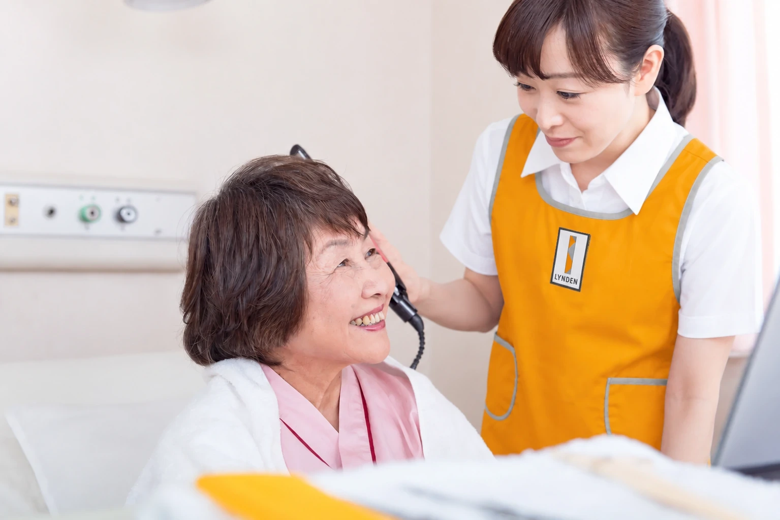 病室のベッドで髪を整えるオレンジ色の制服のスタッフと笑顔の利用者